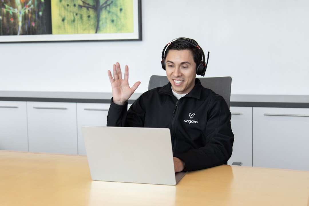 about-01 A customer support representative at Vagaro, smiling and waving, is assisting a client through a headset in an office, offering support.